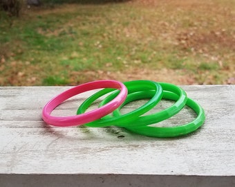 Può includere: Quattro braccialetti colorati. Uno è rosa acceso e gli altri tre sono verdi. I braccialetti sono appoggiati su una superficie di legno bianco usurato. Lo sfondo è una vista sfocata di erba verde e fogliame.