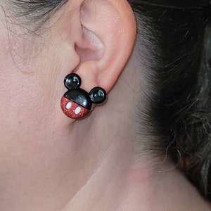 K&ouml;nnte beinhalten: Nahaufnahme eines Ohrs mit einem Mickey Mouse-Ohrstecker. Der Ohrring hat einen schwarzen Kopf mit zwei schwarzen kreisf&ouml;rmigen Ohren, einem roten Glitzergesicht und einem wei&szlig;en Mund. Ein lustiges Accessoire.