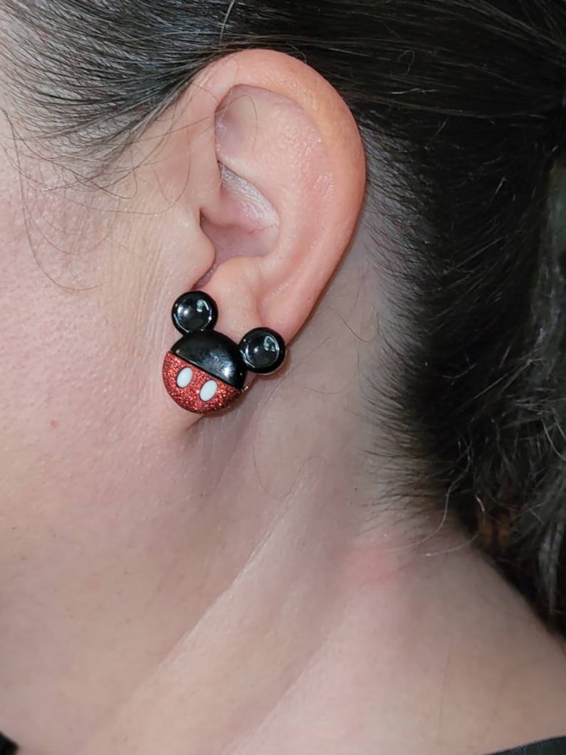 K&ouml;nnte beinhalten: Nahaufnahme eines Ohrs mit einem Mickey Mouse-Ohrstecker. Der Ohrring hat einen schwarzen Kopf mit zwei schwarzen kreisf&ouml;rmigen Ohren, einem roten Glitzergesicht und einem wei&szlig;en Mund. Ein lustiges Accessoire.