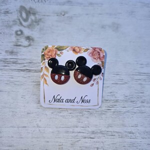 K&ouml;nnte beinhalten: Ohrstecker in Form von Mickey Mouse. Die Ohrringe haben schwarze Ohren und rot glitzernde Gesichter mit wei&szlig;en Akzenten. Sie werden auf einer Karte mit floralen Akzenten und dem Text "Nala and Ness" pr&auml;sentiert.