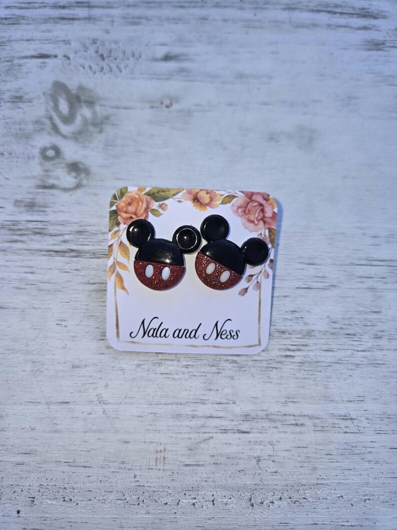 K&ouml;nnte beinhalten: Ohrstecker in Form von Mickey Mouse. Die Ohrringe haben schwarze Ohren und rot glitzernde Gesichter mit wei&szlig;en Akzenten. Sie werden auf einer Karte mit floralen Akzenten und dem Text "Nala and Ness" pr&auml;sentiert.