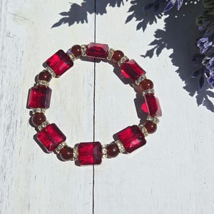 May include: A red and gold bracelet featuring rectangular red glass beads and smaller brown beads. The rectangular beads are accented with small, clear crystals. The bracelet is displayed on a white wooden surface.