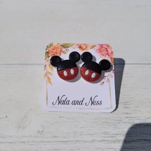K&ouml;nnte beinhalten: Ein Paar Ohrstecker in Form von Mickey Mouse. Die Ohrringe haben ein rotes Glitzergesicht mit wei&szlig;en ovalen Augen und schwarzen Ohren. Sie werden auf einer Blumenkarte mit dem Text "Nala and Ness" pr&auml;sentiert.