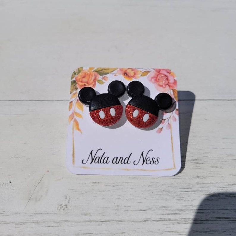 K&ouml;nnte beinhalten: Ein Paar Ohrstecker in Form von Mickey Mouse. Die Ohrringe haben ein rotes Glitzergesicht mit wei&szlig;en ovalen Augen und schwarzen Ohren. Sie werden auf einer Blumenkarte mit dem Text "Nala and Ness" pr&auml;sentiert.