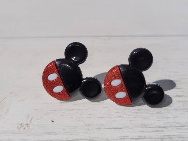 K&ouml;nnte beinhalten: Ein Paar Ohrringe in Form von Mickey Mouse. Jeder Ohrring hat einen schwarzen Kopf mit zwei kleinen schwarzen Ohren und einem roten, glitzernden Gesicht mit zwei wei&szlig;en Ovalen. Die Ohrringe sind vor einem wei&szlig;en Hintergrund platziert.
