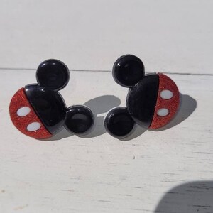 K&ouml;nnte beinhalten: Ein Paar schwarze und rote Ohrringe in Mickey Mouse-Form. Die Ohrringe haben einen schwarzen Kopf und Ohren mit einem roten, glitzernden Gesicht. Zwei wei&szlig;e Kreise befinden sich auf dem roten Gesicht. Die Ohrringe liegen auf einer wei&szlig;en Oberfl&auml;che.