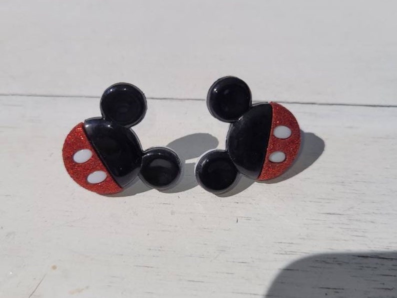 K&ouml;nnte beinhalten: Ein Paar schwarze und rote Ohrringe in Mickey Mouse-Form. Die Ohrringe haben einen schwarzen Kopf und Ohren mit einem roten, glitzernden Gesicht. Zwei wei&szlig;e Kreise befinden sich auf dem roten Gesicht. Die Ohrringe liegen auf einer wei&szlig;en Oberfl&auml;che.