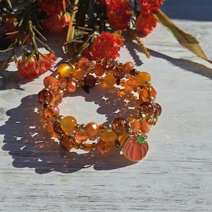 Könnte beinhalten: Ein mehrreihiges Wickelarmband mit einer Vielzahl von orangefarbenen, bernsteinfarbenen und goldfarbenen Perlen in verschiedenen Größen und Texturen. Das Armband enthält einen kleinen, hängenden orangefarbenen Kürbisanhänger.