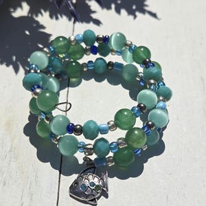 Peut inclure: Un bracelet enroulé avec des perles vertes et bleues, agrémenté d'entretoises argentées et d'un charm en forme de poisson. Le bracelet présente différentes nuances de perles vertes et bleues, créant un effet superposé. Le charm en forme de poisson ajoute une touche décorative.
