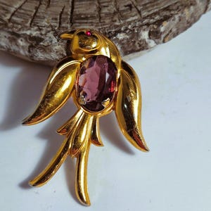 May include: A gold-tone bird brooch with a large, oval-shaped purple rhinestone in the center. The bird has a small red rhinestone for an eye.