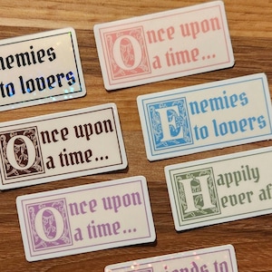 May include: Rectangular stickers with text in various colors and fonts. Phrases include "Enemies to lovers," "Once upon a time...", "Happily ever after," and "Friends to lovers." The stickers have a white background with decorative borders.