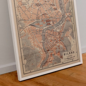 May include: A vintage map of Bilbao, Spain, printed in black and white with red accents. The map shows the city's streets, buildings, and river. The title "Bilbao" is printed in the lower right corner, along with the scale "1:15,000".