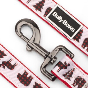 以下が含まれることがあります： 白い背景と赤い縁取りの犬用リード。リードには、黒と赤のチェック柄のクマ、木、ヘラジカの繰り返し模様があります。リードには黒い金属製の留め金と、「Bully Bows」と書かれた白黒のラベルが付いています。