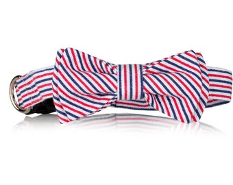 May include: A red, white, and blue striped bow tie collar for a pet. The collar is made of fabric and has a metal buckle.
