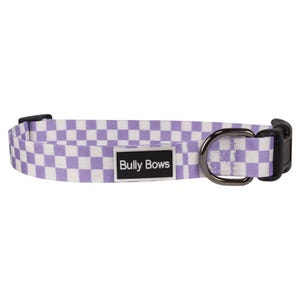 May include: A dog collar with a purple and white checkered pattern. The collar has a black plastic buckle and a metal D-ring. A rectangular label with the text "Bully Bows" is centered on the collar.