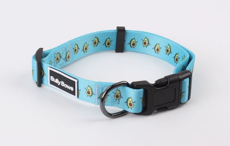 May include: A light blue dog collar with a repeating pattern of cartoon avocados. The collar has a black buckle and a black metal D-ring. The collar has a white label that says "Bully Bows".