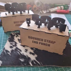 Op de afbeelding: Twee houten dozen met zwarte metalen ponsen. Op één doos staat "BELT END PUNCH" en op de andere "ROUNDED STRAP END PUNCH". De dozen staan op een oppervlak met koeienvelpatroon en een groene snijmat.