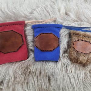 May include: Three small pouches in red, blue, and brown fur. Each pouch has a brown leather patch with black stitching. The pouches have drawstrings in matching colors. The background is a fluffy, light gray surface.