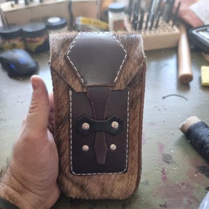 May include: A brown leather phone case with a white fur exterior. The case has a flap with a brown leather strap and silver rivets.