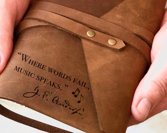 May include: A brown leather journal with a quote engraved on the front: "Where words fail, music speaks." The quote is attributed to "F.F. Schubert".