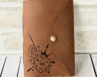 May include: A brown leather journal with a black ink splatter design and a gold button closure. The design features a set of dice, including a 20-sided die.