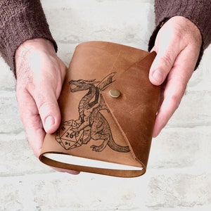 May include: A brown leather journal featuring a dragon and a twenty-sided die design. The journal has a flap closure and a brass button.