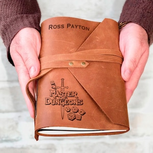 May include: A brown leather-bound journal with a strap closure. The cover is embossed with the text "Master of Dungeons" and a sword and dice graphic. The name "Ross Payton" is also embossed on the cover.
