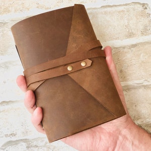 May include: A brown leather journal with a wrap-around closure and brass accents. The journal is held in a hand.