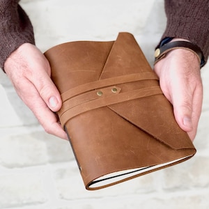 May include: A brown leather journal with a folded cover and a strap closure. The journal has brass accents and is held in a person's hands.