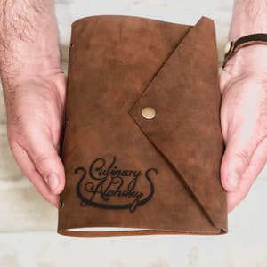 Puede incluir: Un diario de cuero marr&oacute;n con cierre de solapa y bot&oacute;n de lat&oacute;n. El diario est&aacute; estampado con el texto "Culinary Alchemy" en una fuente cursiva.
