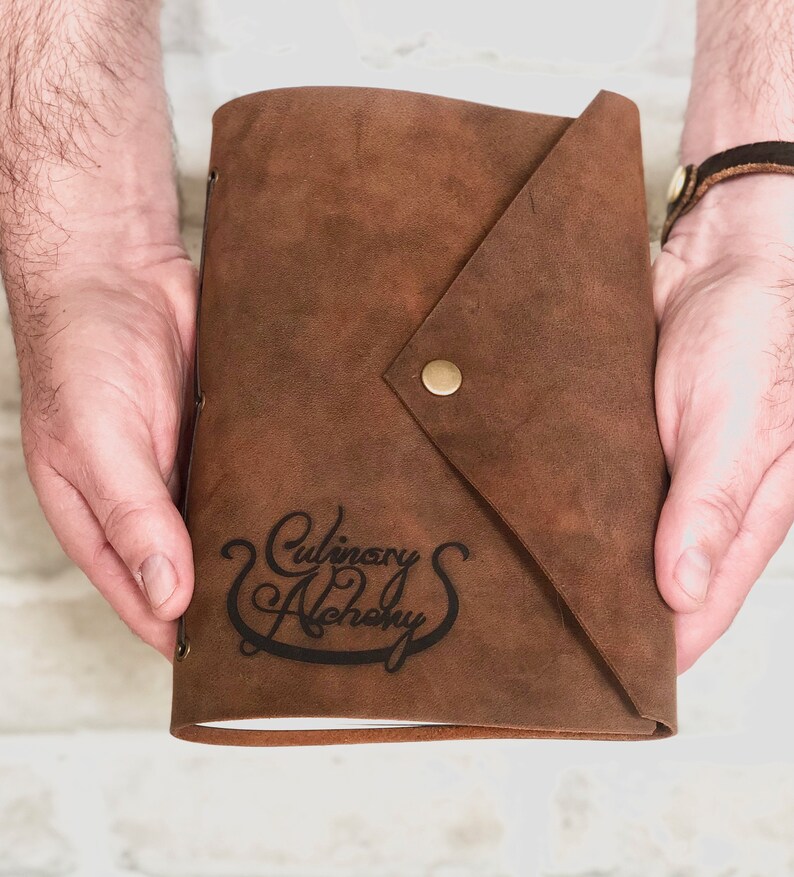 Puede incluir: Un diario de cuero marr&oacute;n con cierre de solapa y bot&oacute;n de lat&oacute;n. El diario est&aacute; estampado con el texto "Culinary Alchemy" en una fuente cursiva.
