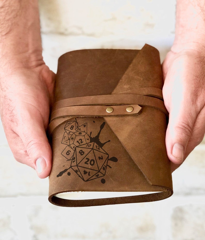 May include: A brown leather journal with a design of dice and a paint splatter. The journal has a strap closure with brass accents.