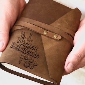 May include: A brown leather journal with a strap closure and a laser engraved design featuring the text "Master of Dungeons" with a sword and dice.