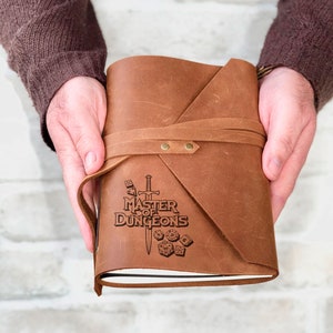 May include: A brown leather-bound journal with a strap closure. The cover is embossed with the text "Master of Dungeons" and a sword and dice design.