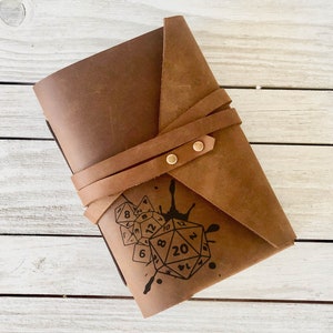 May include: A brown leather journal with a strap closure and a black ink design of dice and a splatter pattern. The journal is closed and the strap is wrapped around it.