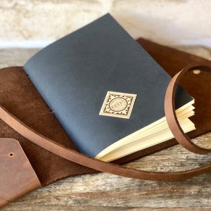 May include: A brown leather journal with a black cover and a small, cream-colored label that says "KEEP" in a decorative font. The journal is closed and has a brown leather strap.