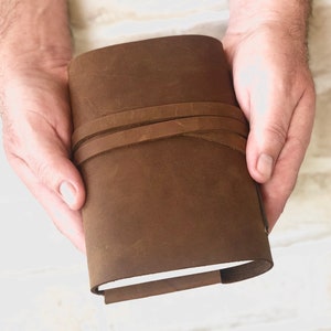 May include: A brown leather journal with a strap closure. The journal is closed and held in a person's hands.