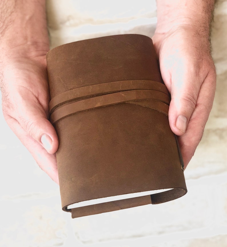 May include: A brown leather journal with a strap closure. The journal is closed and held in a person's hands.