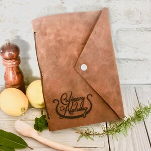 Puede incluir: Un cuaderno de recetas de cuero marr&oacute;n con el texto "Culinary Alchemy" grabado en la portada. El cuaderno est&aacute; sobre una superficie de madera con una cuchara de madera, limones y hierbas.