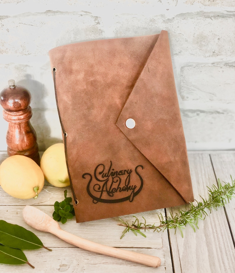 Puede incluir: Un cuaderno de recetas de cuero marr&oacute;n con el texto "Culinary Alchemy" grabado en la portada. El cuaderno est&aacute; sobre una superficie de madera con una cuchara de madera, limones y hierbas.