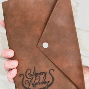 Puede incluir: Un diario de cuero marr&oacute;n con cierre de solapa y bot&oacute;n met&aacute;lico. El diario tiene las palabras "Culinary Alchemy" grabadas en la parte delantera.