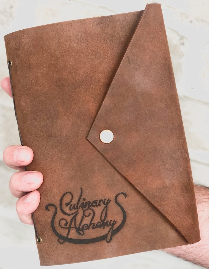 Puede incluir: Un diario de cuero marr&oacute;n con cierre de solapa y bot&oacute;n met&aacute;lico. El diario tiene las palabras "Culinary Alchemy" grabadas en la parte delantera.