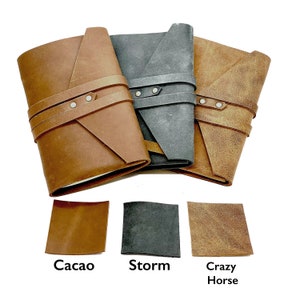 May include: Three leather-bound journals in different colors: brown, black, and tan. Each journal has a leather strap with metal snaps and a folded flap design. The journals are arranged in a row, with the brown journal on the left, the black journal in the middle, and the tan journal on the right. Below the journals are three leather swatches labeled "Cacao", "Storm", and "Crazy Horse".