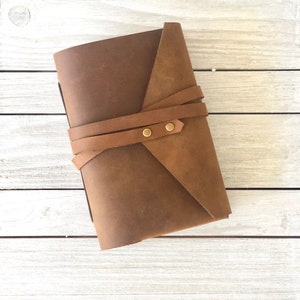 May include: A brown leather journal with a strap closure and brass accents. The journal is closed and lying flat on a white wooden surface.