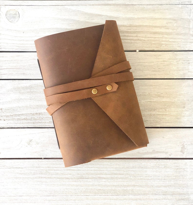 May include: A brown leather journal with a strap closure and brass accents. The journal is closed and lying flat on a white wooden surface.