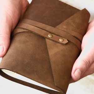 May include: A brown leather journal with a flap closure and a strap with brass accents. The journal is held in a person's hands.
