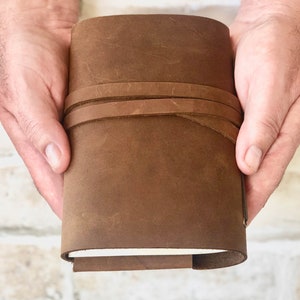 May include: A brown leather journal with a strap closure. The journal is closed and held in a person's hands.