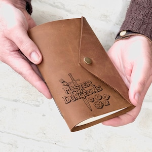May include: A brown leather journal with a flap closure and a brass button. The cover is embossed with the words "MASTER OF DUNGEONS" along with dice and a sword. The journal is held open, revealing the blank pages.