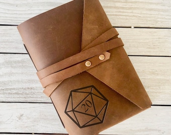 May include: A brown leather journal with a strap closure and a D20 dice design engraved on the front.