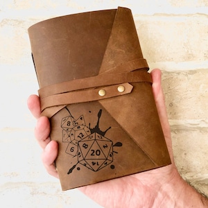May include: A brown leather journal with a strap closure and a design of dice and ink splatters. The journal has a vintage look and feel.
